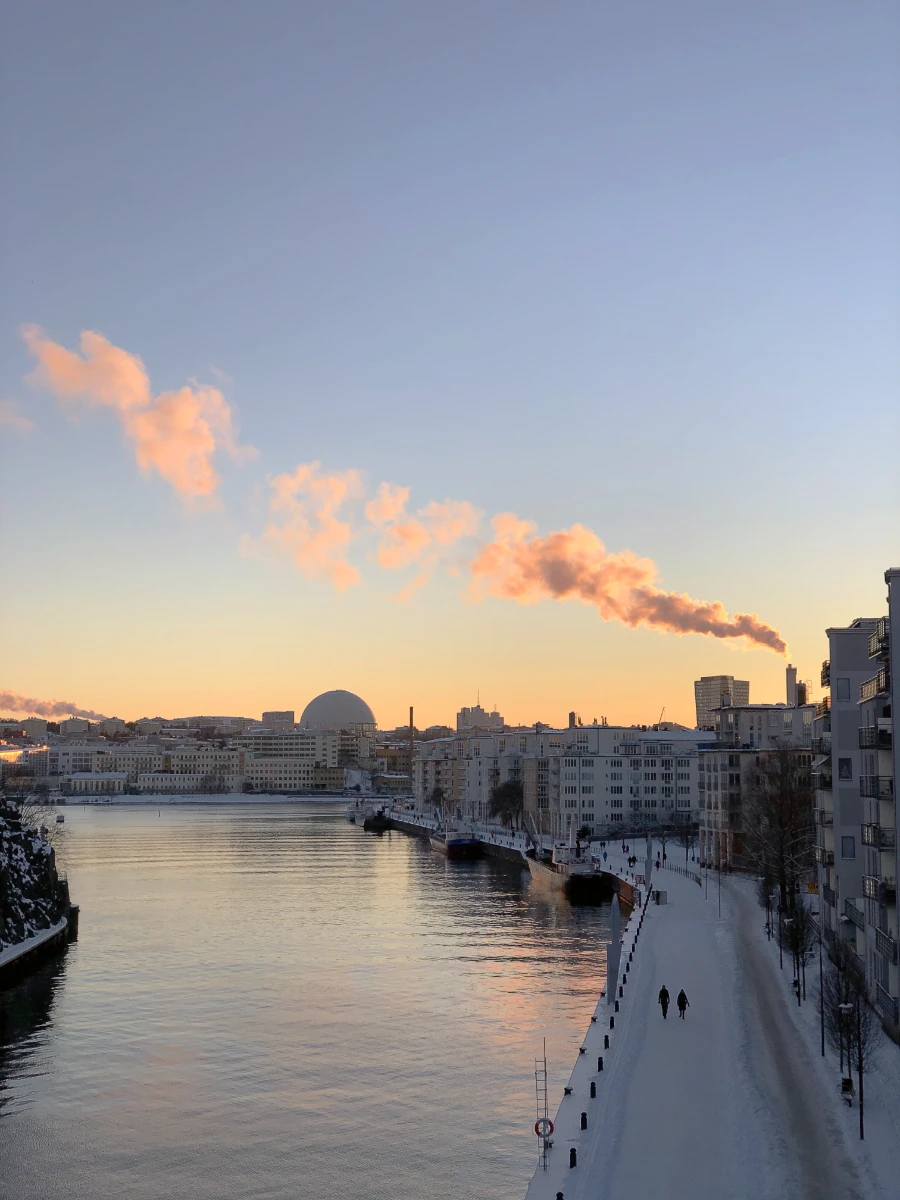 Looking at Hammarby from bridge. Snow covered sidewalks. Smoke from smokestack illuminated by setting sun.