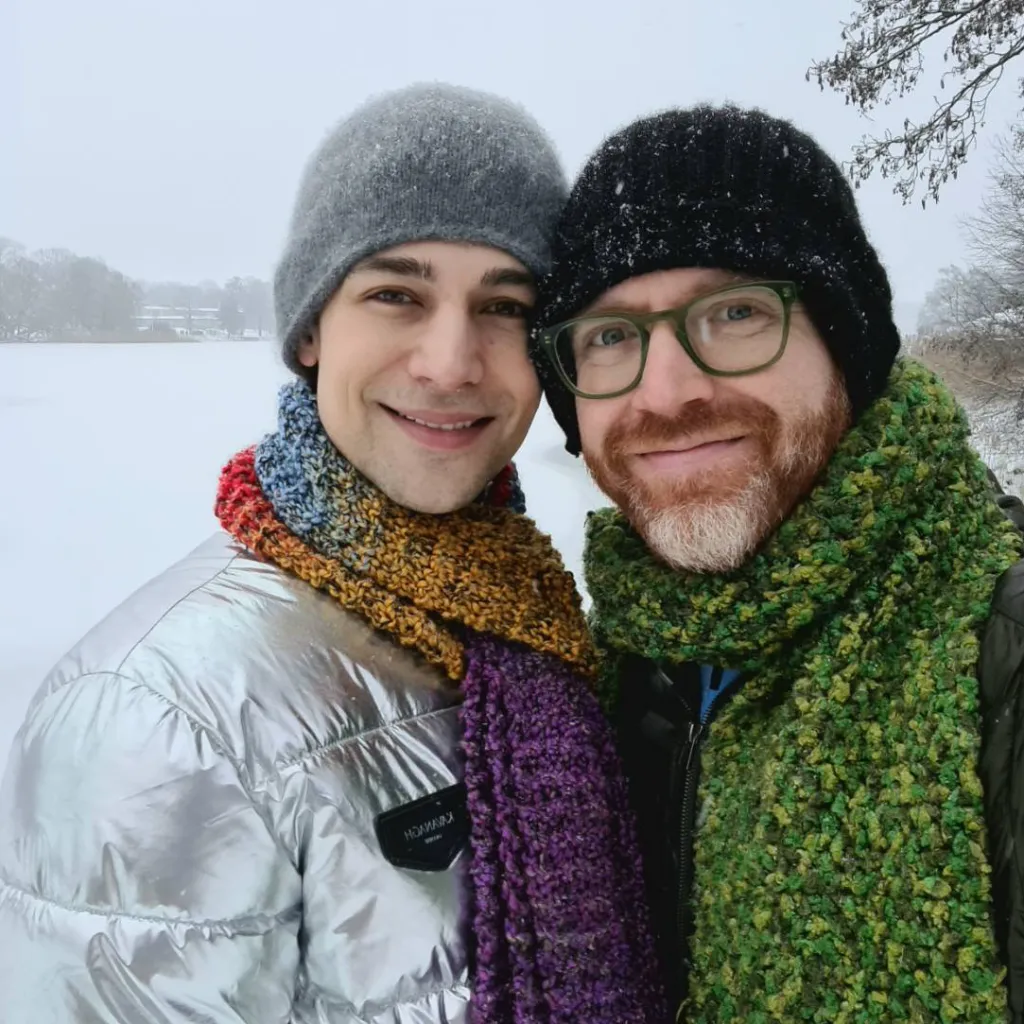 Jeremiah and Arthur outside in snow. Jeremiah in silver coat and rainbow scarf. Arthur in black coat with green scarf.