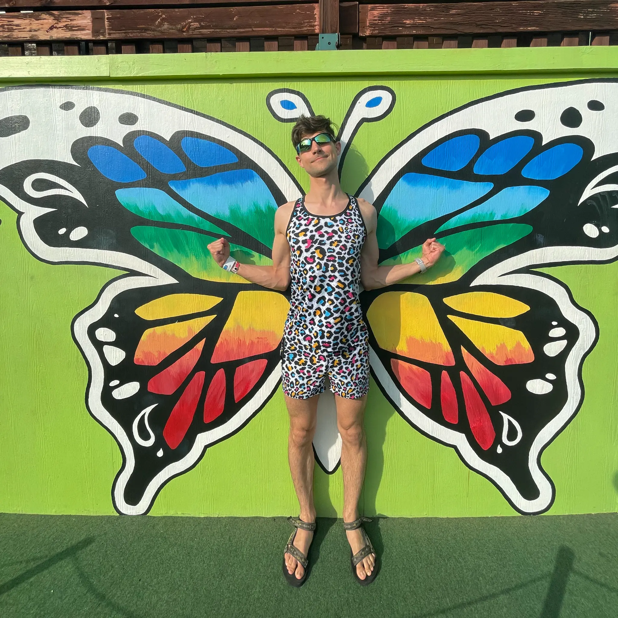 Jeremiah in rainbow cheetah print swim trucks and tanktop standing in front of a butterfly wing mural