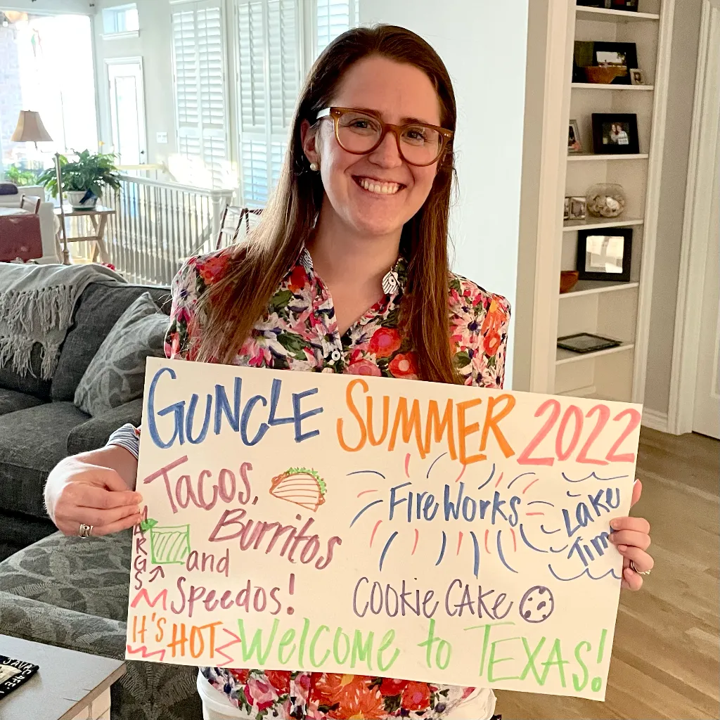 Adrienne holding sign: Guncle Summer 2022. Tacos, margs, speedos, cookie cake, fireworks, lake time. Welcome to Texas!