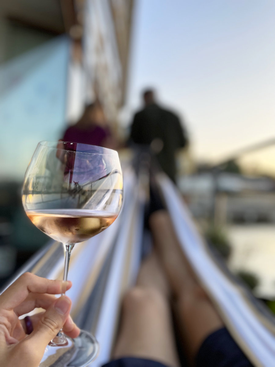Jeremiah holding a glass of wine while laying in a hammock. Karin and Arthur working on grill in background.