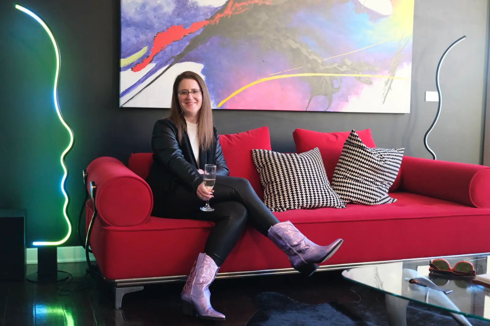 Adrienne dressed in black leather pants and jacket with purple cowgirl boots sitting cross-legged on a red couch
