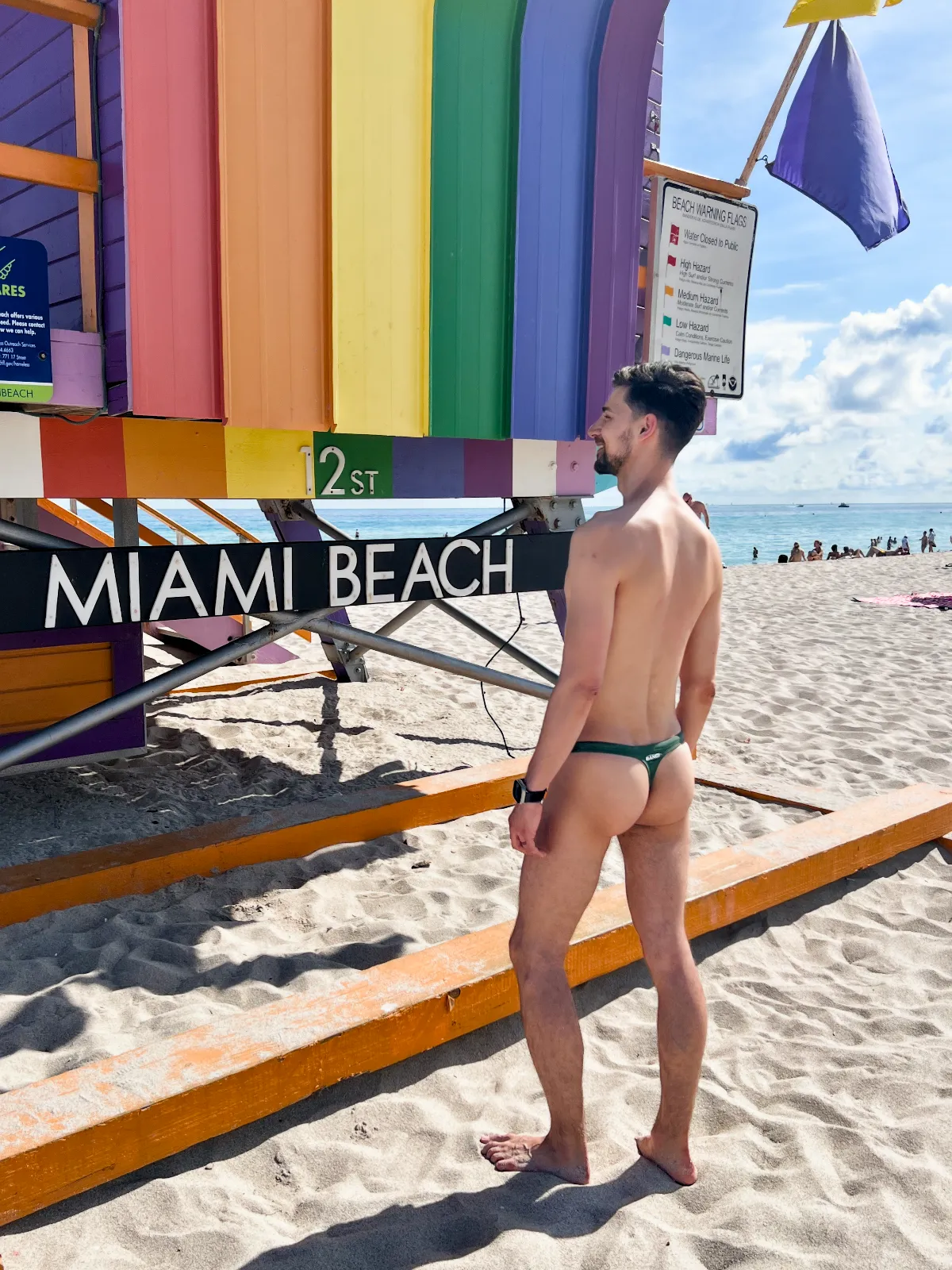 Jeremiah wearing a thong facing the rainbow striped lifeguard house