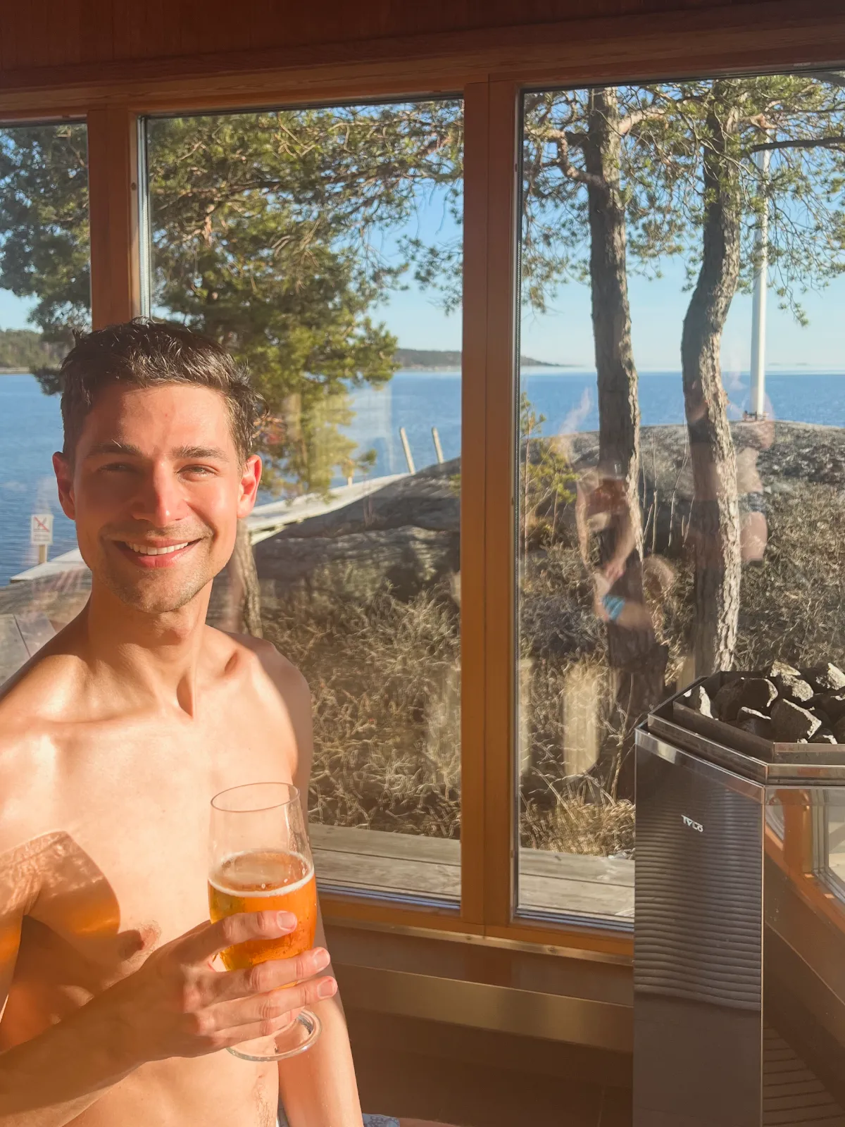 Jeremiah holding a beer in a sauna with a view of the archipelago out of the window
