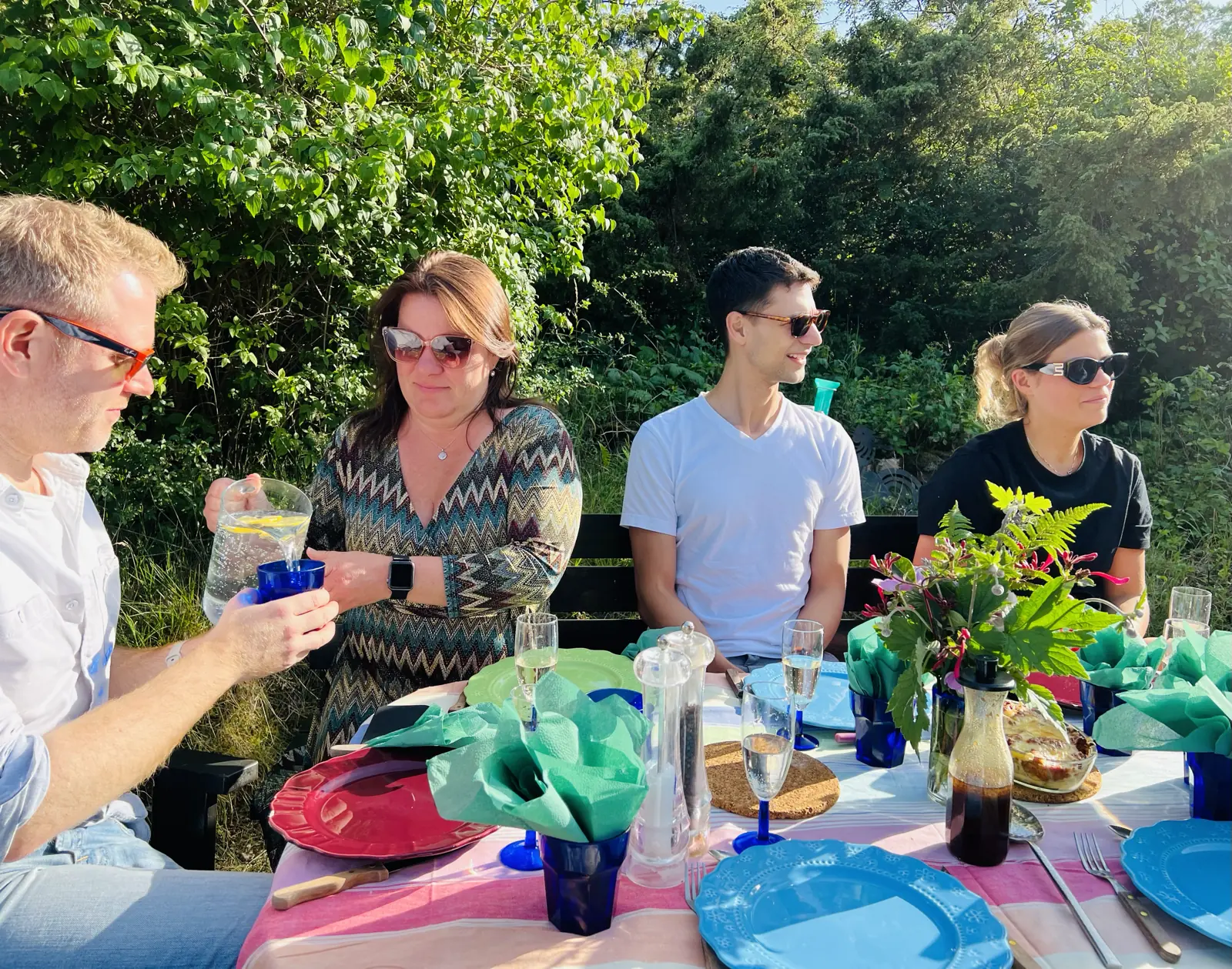 Arthur, Karin, Jeremiah, Vendela sitting at a table outside with brightly colored plates and fresh flowers. It’s sunny and everyone is wearing sunglasses.