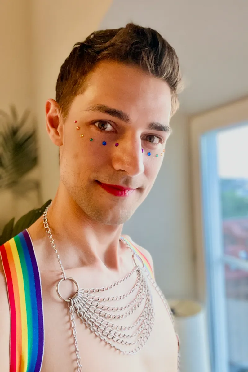 Jeremiah shirtless wearing rainbow harness, metal chain necklace, red lipstick, and rainbow jewels under eyes