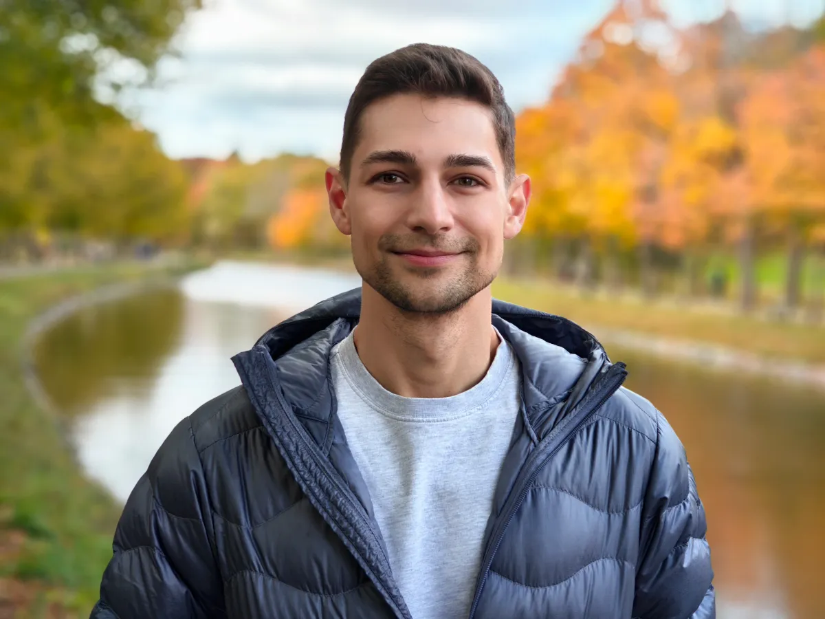 Jeremiah standing in a park with tree changing colors in the fall alongside a river