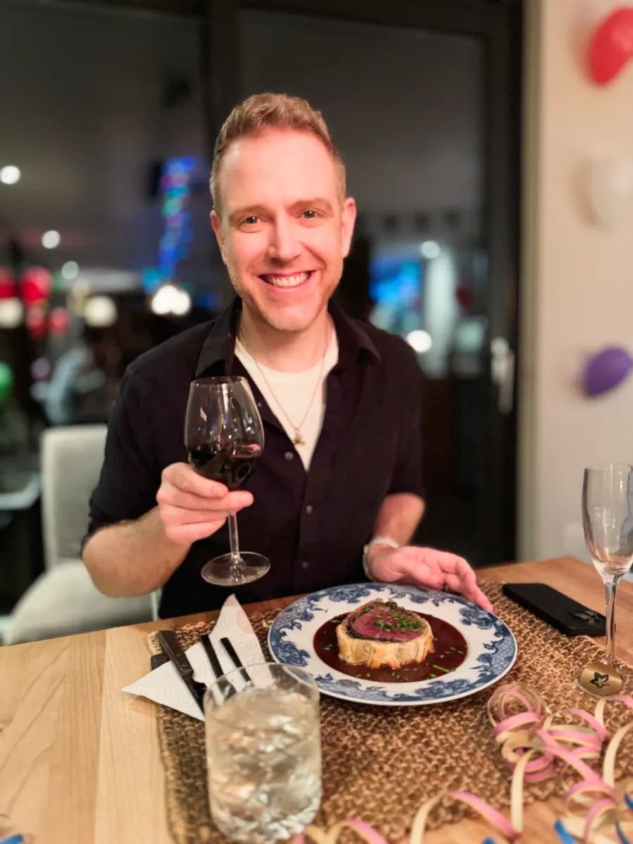 Arthur smiling holding glass of wine with a plate of beef wellington in front of him