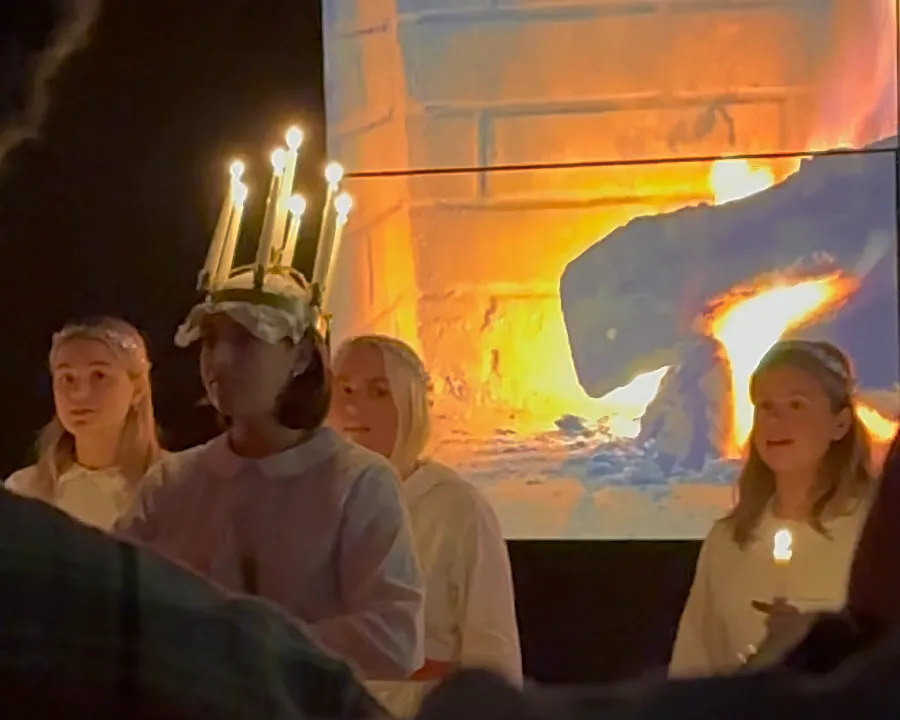 Lucia choir. Young woman wears crown with lighted candles. 3 women behind her. Standing in front of a projection of a fireplace.
